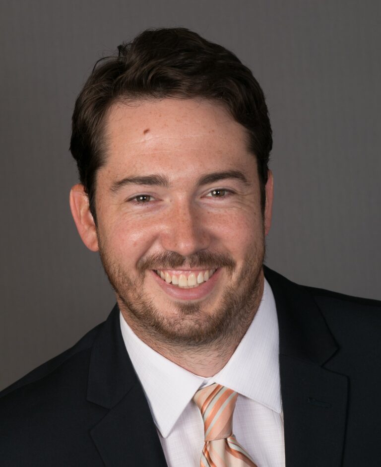 Ben Clark is wearing a suit and tie, smiling at the camera with a dark grey background behind him.