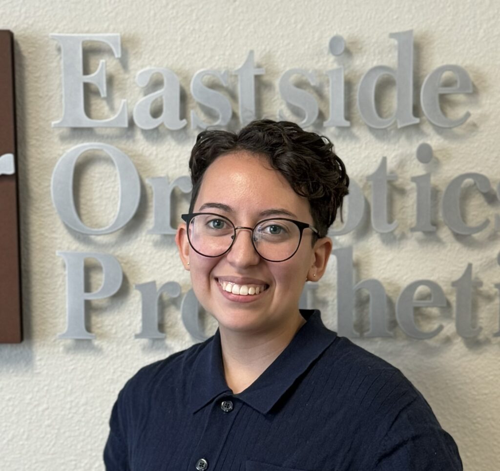 Head shot of Noelle wearing black wire glasses and a navy blue polio. Noelle is smiling at the camera while standing infront of the Eastside O and P sign.